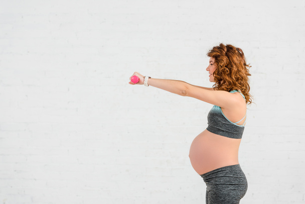 Side view of smiling pregnant woman training with dumbbells on white background - Photo, Image