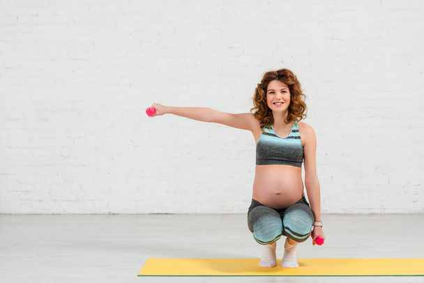 Femme enceinte souriant à la caméra et faisant de l'exercice avec des haltères sur tapis de fitness à la maison
 - Photo, image