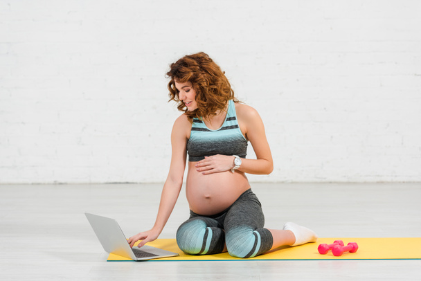 Χαμογελώντας έγκυος γυναίκα χρησιμοποιώντας φορητό υπολογιστή στο στρώμα γυμναστικής κοντά αλτήρες στο στρώμα γυμναστικής στο σπίτι  - Φωτογραφία, εικόνα