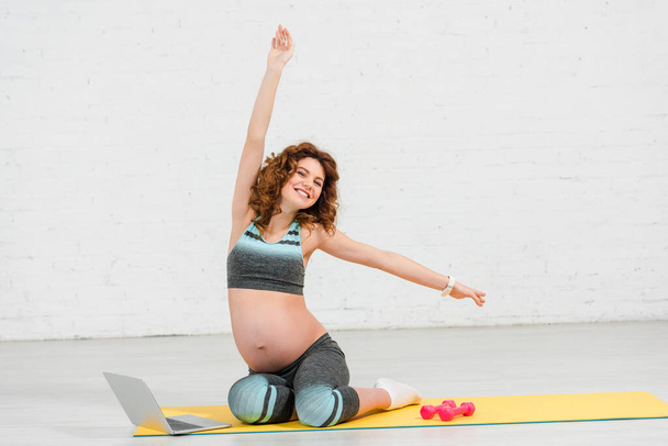 Schwangere lächelt beim Training neben Laptop und Hanteln auf Fitnessmatte in die Kamera - Foto, Bild