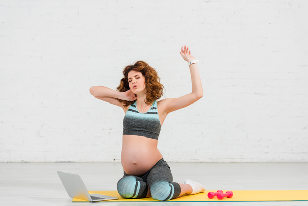 Zwangere vrouw raakt nek tijdens de training in de buurt laptop en halters op fitness mat - Foto, afbeelding