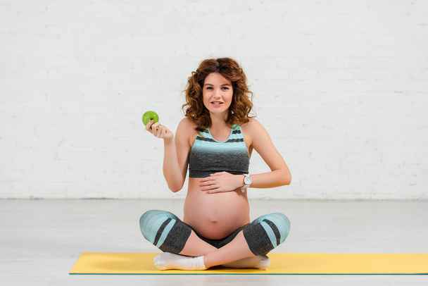 Pregnant woman holding apple and smiling at camera on fitness mat - Fotoğraf, Görsel