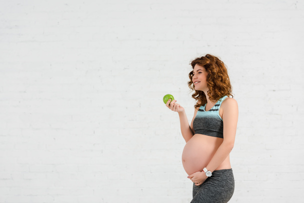 Вид сбоку улыбающейся беременной женщины, держащей яблоко на белом фоне
 - Фото, изображение