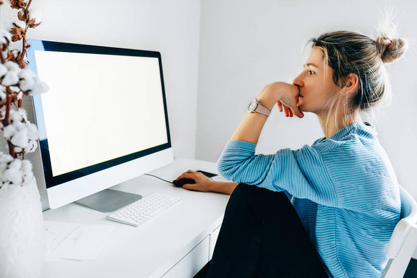 Side view image of freelancer businesswoman using a desktop computer in an office. Young blonde woman sits indoors at home working on the computer with a blank screen for your text message or content. - Фото, зображення
