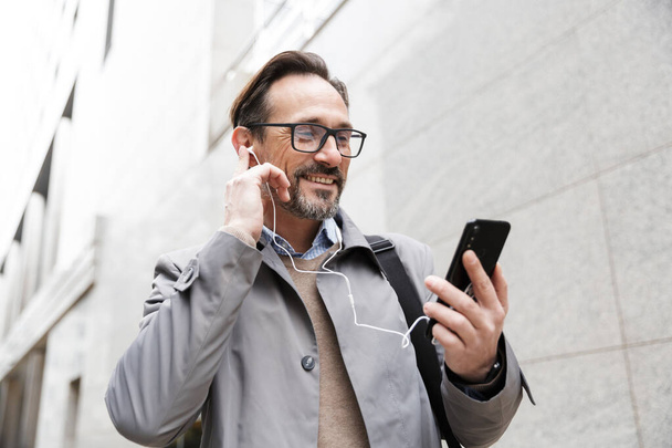 Image of joyful businessman using cellphone and earphones - Фото, изображение