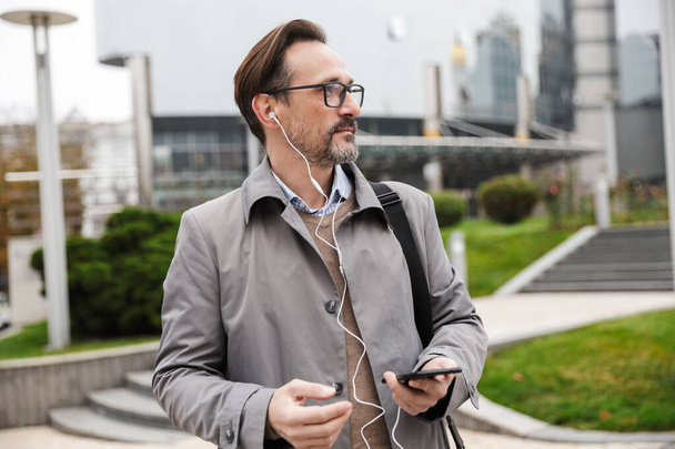 Image of confident businessman using cellphone and earphones - Foto, afbeelding