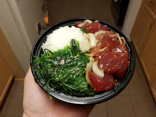 hand holding bowl of raw tuna and green seaweed and rice in kitchen - Foto, afbeelding