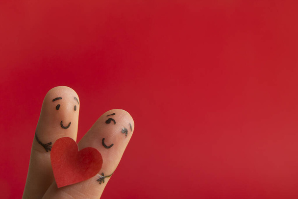 День Святого Валентина в любви концепции. Раскрашенные счастливые смешные пальцы улыбаются держа красное сердце на красном фоне с копией места для текста объявления
 - Фото, изображение