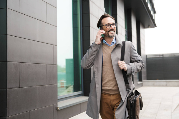 Image of mature smiling businessman talking on cellphone while walking - Foto, Imagen