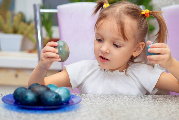 Little charming European girl with two ponytails sits at a table and plays with painted Easter eggs. - Photo, Image