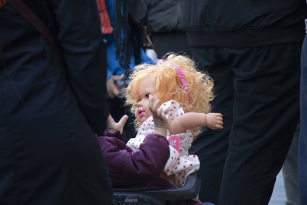 Дитина в колясці фотографується, граючи зі своєю лялькою. Навколо багато людей
. - Фото, зображення