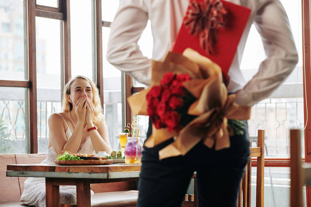Horizontal shot of unrecognizable young man showing up for date in cafe with rose bouquet and box of chocolate for young woman - Fotoğraf, Görsel