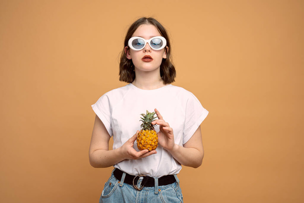 Beyaz güneş gözlüklü şık bir bayan ananasları elinde tutuyor ve kameraya bakıyor. - Fotoğraf, Görsel
