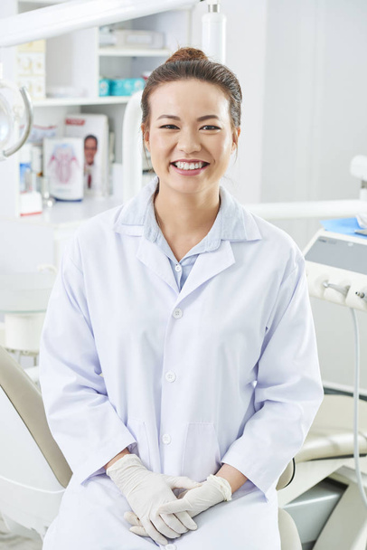 Vertical medium long shot of attractive Asian female dentist sitting in her office looking at camera, smiling - Photo, Image