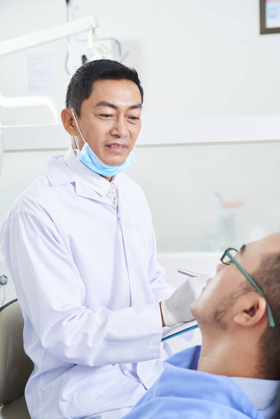 Vertical portrait of Asian man wearing mask and white coat consulting patient in dental clinic office - Photo, Image