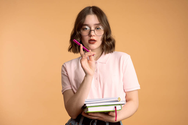 Image of successful female writer or journalist, makes notes in diary, holds pencil near mouth, wears round glasses, has dark hair, isolated over orange studio wall. - Fotó, kép