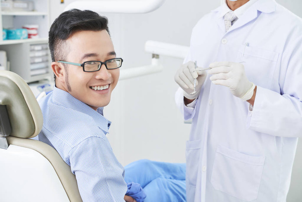 Poziome ujęcie wesołego azjatyckiego pacjenta kliniki dentystycznej siedzącego na krześle uśmiechniętego, nierozpoznawalnego dentysty stojącego obok niego - Zdjęcie, obraz
