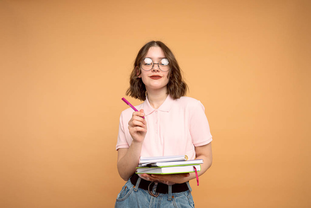 Στιγμιότυπο του θετικού μαθητή κορίτσι φορώντας γυαλιά, δείχνοντας προς τα πάνω με στυλό, κρατώντας σημειωματάριο, αναζητούν ενθουσιασμένοι, απομονώνονται σε πορτοκαλί φόντο - Φωτογραφία, εικόνα