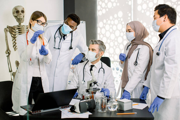 Group of five multiethnic laboratory scientists in lab coats, masks and gloves, working at lab with test tubes, microscope, flasks, laptop, developing new substance, medicine or vaccine - Foto, afbeelding