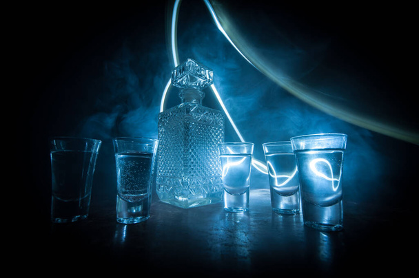 A beautiful carafe with vodka stands on a wooden table with glasses. Dramatic toned foggy background with light. Selective focus. Club drink concept. - 写真・画像