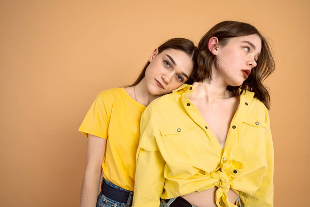 Two young brunette girls in yellow clothes over orange background, european woman leaned on her friend shoulder and looking at the camera - 写真・画像