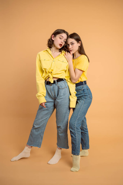 Stylové ženy v modrých džínách a žlutých košilích stojící vedle sebe a dívající se do kamery, izolované přes oranžové pozadí - Fotografie, Obrázek
