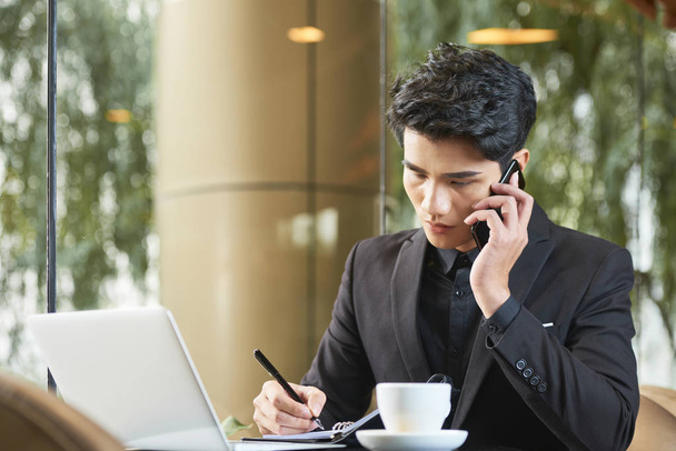 Красивый азиатский бизнесмен в стильной одежде сидит в современном кафе и разговаривает по телефону и делает заметки
 - Фото, изображение