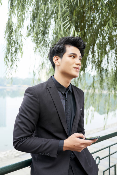 Retrato vertical medio de un hombre asiático guapo con un traje elegante que sostiene el teléfono inteligente pensando en algo
 - Foto, Imagen