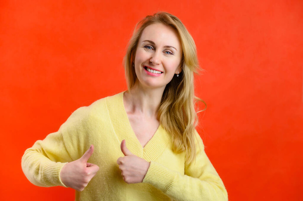 Pretty blonde 40 years old in a yellow sweater shows ok fingers joyfully on a red background - Foto, afbeelding