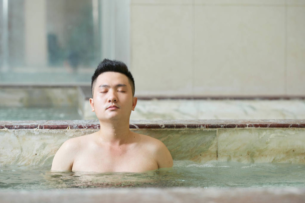 Joven hombre asiático relajante con los ojos cerrados disfrutando de la bañera de hidromasaje en el moderno salón de spa, tiro horizontal - Foto, imagen
