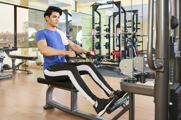 Horizontal full shot of handsome young Asian man doing exercise using fitness machine in spa resort hotel gym - Photo, Image