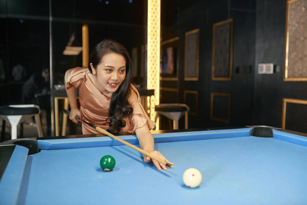Horizontal shot of cheerful young Asian woman entertaining herself playing billiard in hotel lounge - Photo, Image