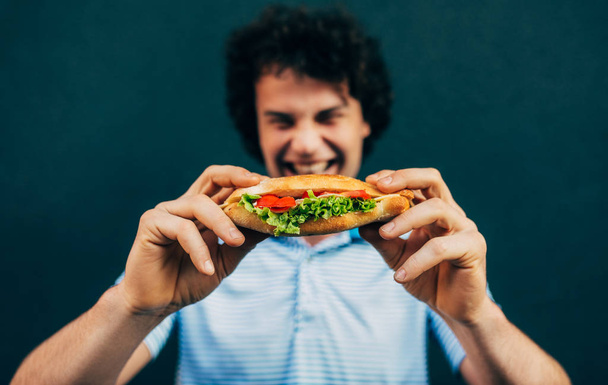 Young man eating a cheeseburger. Happy man in a fast-food restaurant eating a hamburger outdoors. Man with curly hair having street food and eat a burger. - Fotó, kép