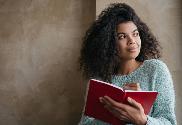 Hermosa mujer afroamericana sosteniendo un libro rojo, mirando a la ventana y sonriendo. Estudiante universitario estudiando, aprendiendo idiomas, sentado en la biblioteca. Retrato de un joven escritor pensativo tomando notas
 - Foto, Imagen