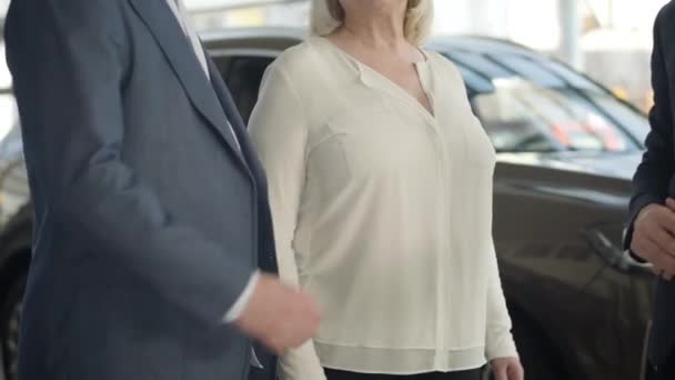 Two unrecognizable Caucasian men in suits shaking hands in front of mature blond woman standing in car dealership. Clients making sale and purchase agreement in showroom. Auto industry, success. - Filmagem, Vídeo