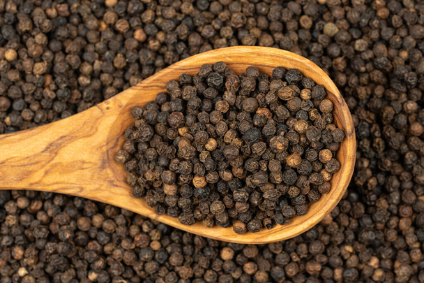 Spice background, Background made of many whole black peppercorns and a cooking spoon made of olive wood also filled with peppercorns , extreme close up, top view - Foto, afbeelding