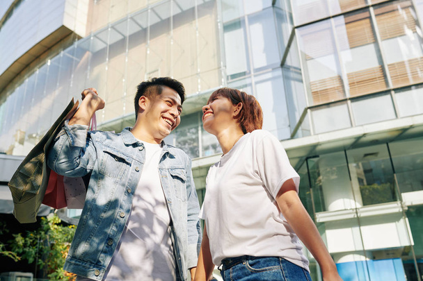 Feliz asiático hombre y mujer de pie juntos contra el centro comercial sosteniendo bolsas de compras sonriendo
 - Foto, imagen