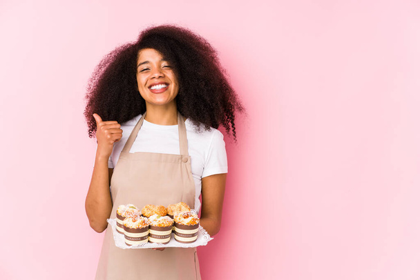 Joven pastelera afro sosteniendo una magdalenas aisladaJoven panadera afro sonriendo y levantando el pulgar hacia arriba
 - Foto, Imagen