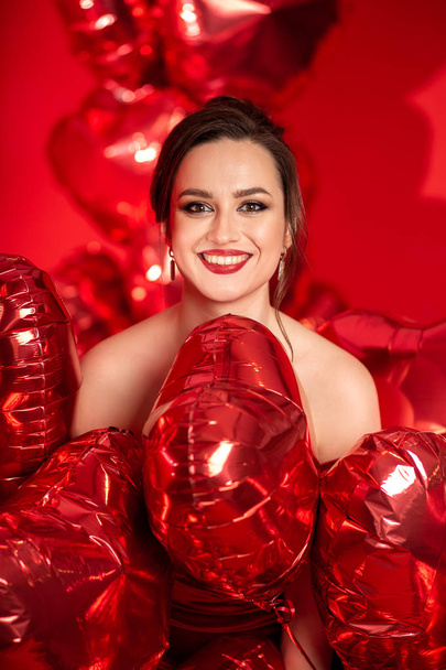 Beautiful young woman in red evening dress posing over red background with big heart shape balloons.  - Fotografie, Obrázek