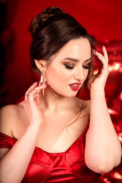 Beautiful young woman in red evening dress posing over red background with big heart shape balloons.  - Fotó, kép