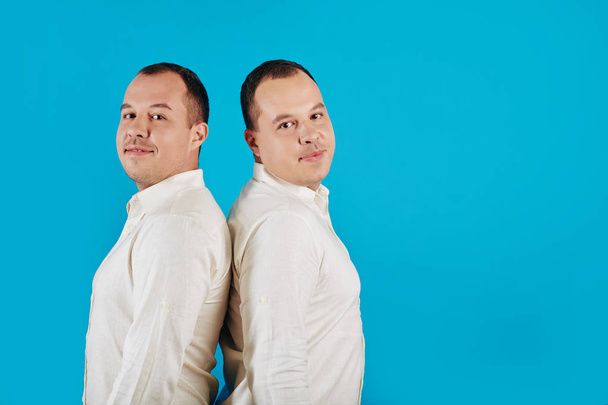 Horizontal studio portrait shot of two twin brothers standing back to back together against blue background looking at camera - Φωτογραφία, εικόνα