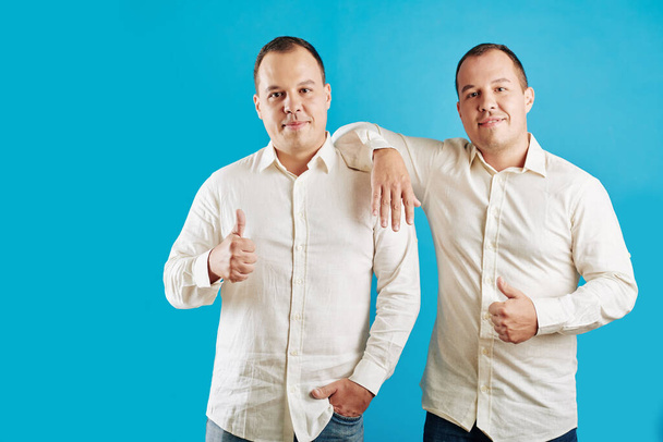 Dwa identyczne samce bliźniaki ubrane w białe koszule stojące na niebieskim tle patrzące na kamerę z kciukami w górze - Zdjęcie, obraz