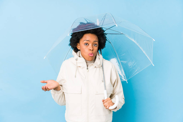 Joven mujer afroamericana sosteniendo un paraguas aislado encoge hombros y los ojos abiertos confundido. - Foto, Imagen