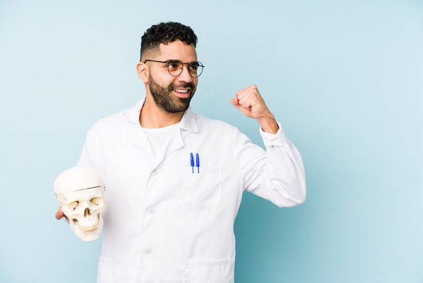 Young doctor latin man holding a skull isolated raising fist after a victory, winner concept. - Photo, Image