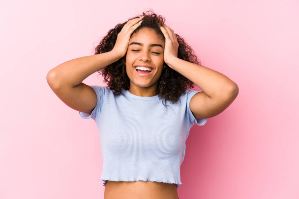 Jonge Afro-Amerikaanse vrouw tegen een roze achtergrond lacht vreugdevol handen op het hoofd. Geluksconcept. - Foto, afbeelding