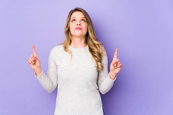 Young caucasian woman isolated on purple background pointing upside with opened mouth. - Photo, image