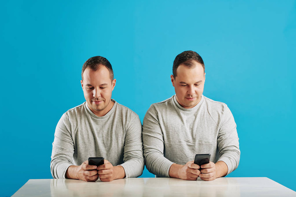 Horizontale shot van tweelingbroers zitten aan tafel kijken naar iets op hun smartphones, heldere blauwe achtergrond - Foto, afbeelding