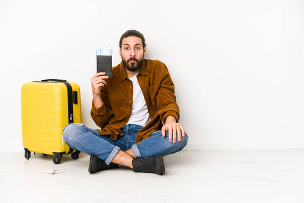 Joven hombre caucásico sentado sosteniendo un pasaporte y una maleta aislado encoge los hombros y los ojos abiertos confundido
. - Foto, Imagen