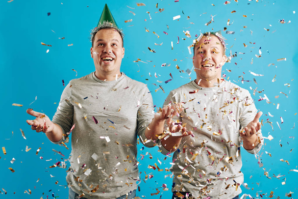 Retrato de estudio horizontal de hermanos gemelos adultos jóvenes celebrando su cumpleaños con confeti, fondo azul
 - Foto, imagen