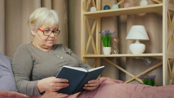 An elderly woman with glasses reads a book in her room - Кадры, видео